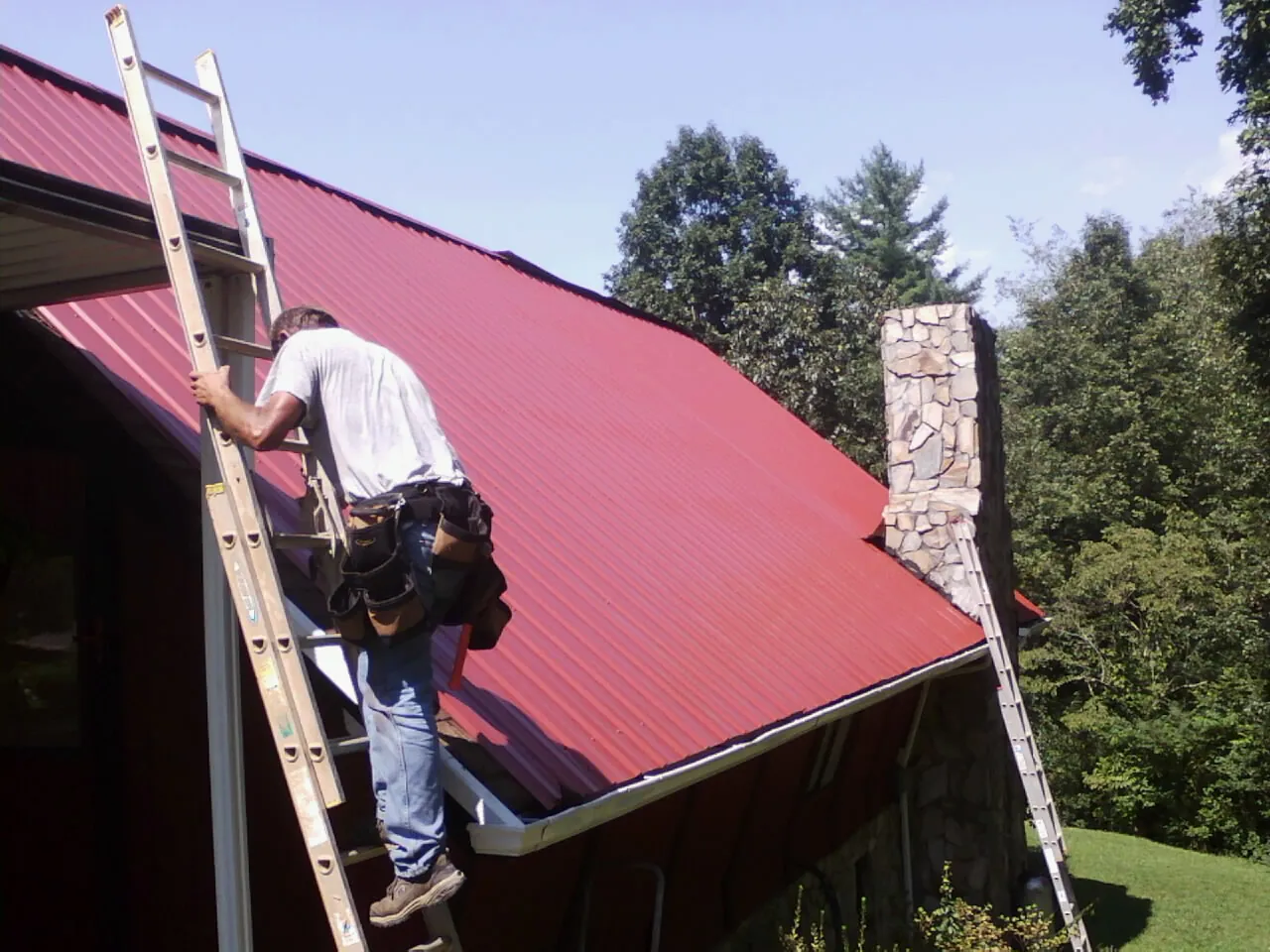 Roofer on a ladder beside a red metal roof and stone chimney — Soffit & Fascia Repair by Roofing Brothers in Clarks Summit